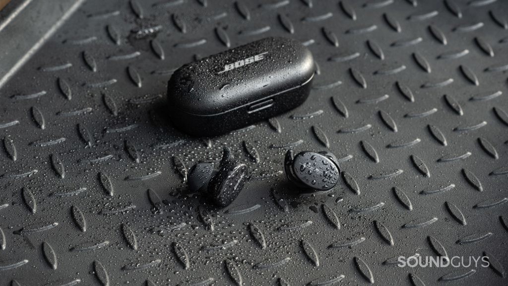 The Bose Sport Earbuds true wireless workout earbuds sit outside of the closed charging case, all objects are covered in sprinkles of water.