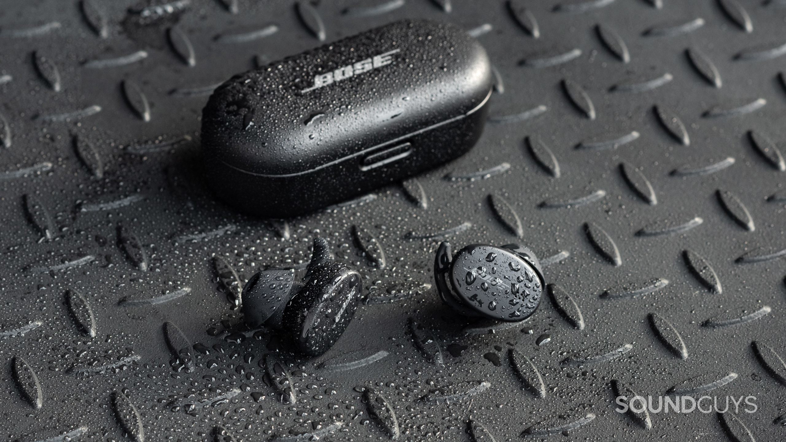 The Bose Sport Earbuds true wireless workout earbuds sit outside of the closed charging case, all objects are covered in sprinkles of water.