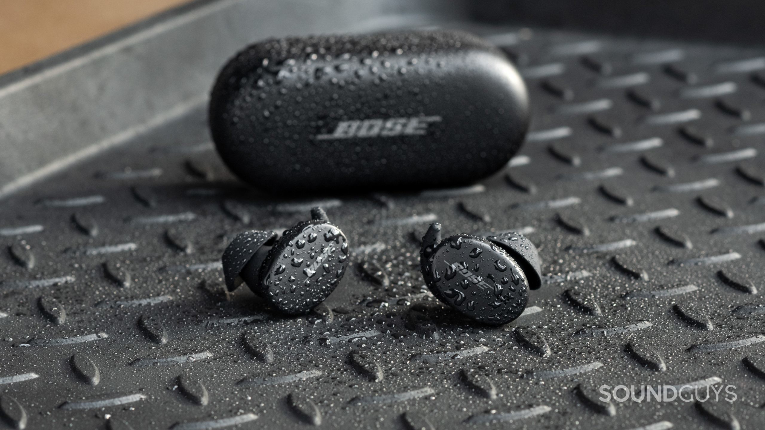 The Bose Sport Earbuds true wireless workout earbuds sit outside of the closed charging case, all objects are covered in sprinkles of water.