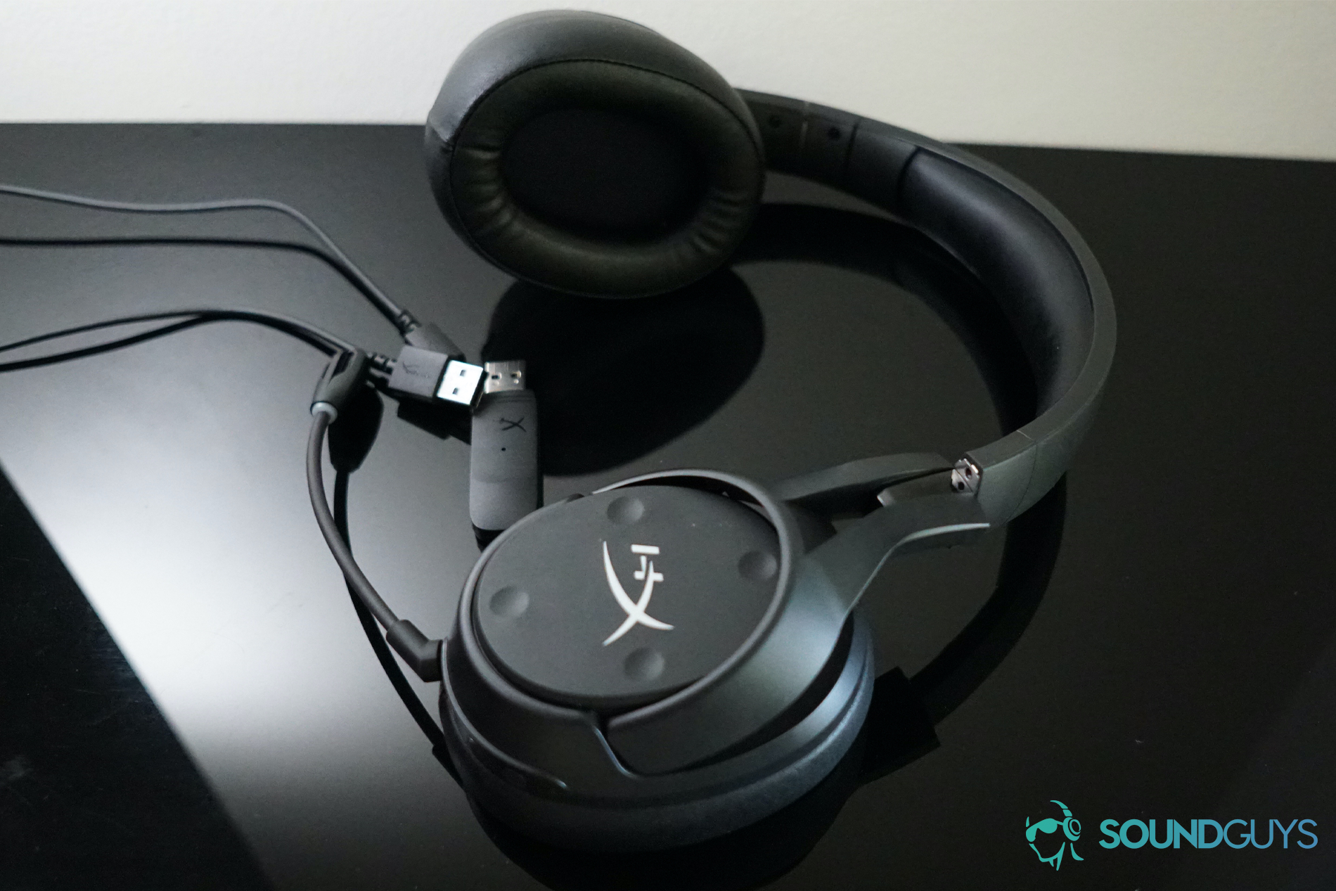 The HyperX Cloud Flight S lays on a reflective surface next to its charging cord and USB dongle.