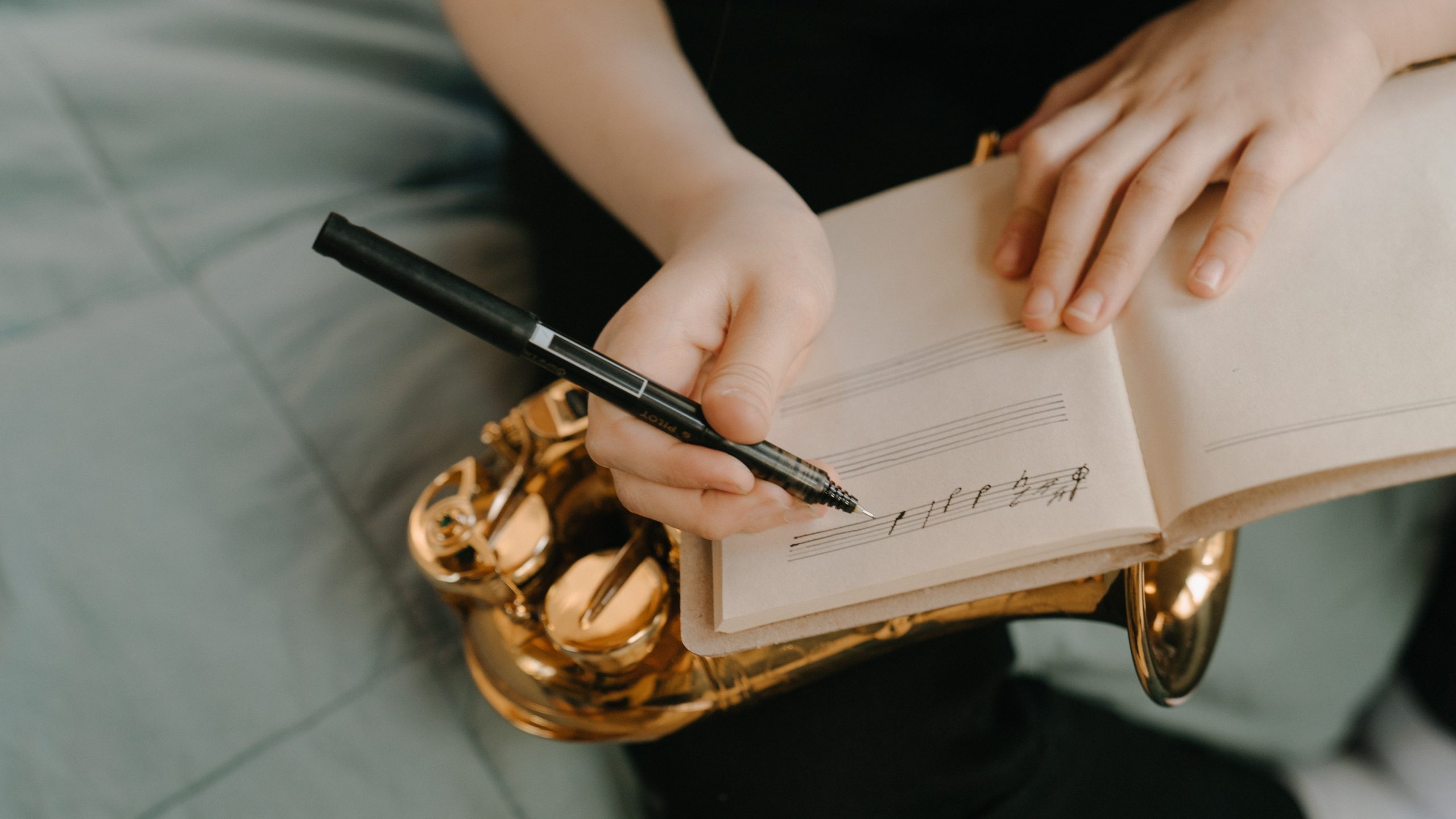 A notebook with music notation being written in it with a pen. Below the notebook is a saxophone resting on a leg. The top of a green duvet is visible.