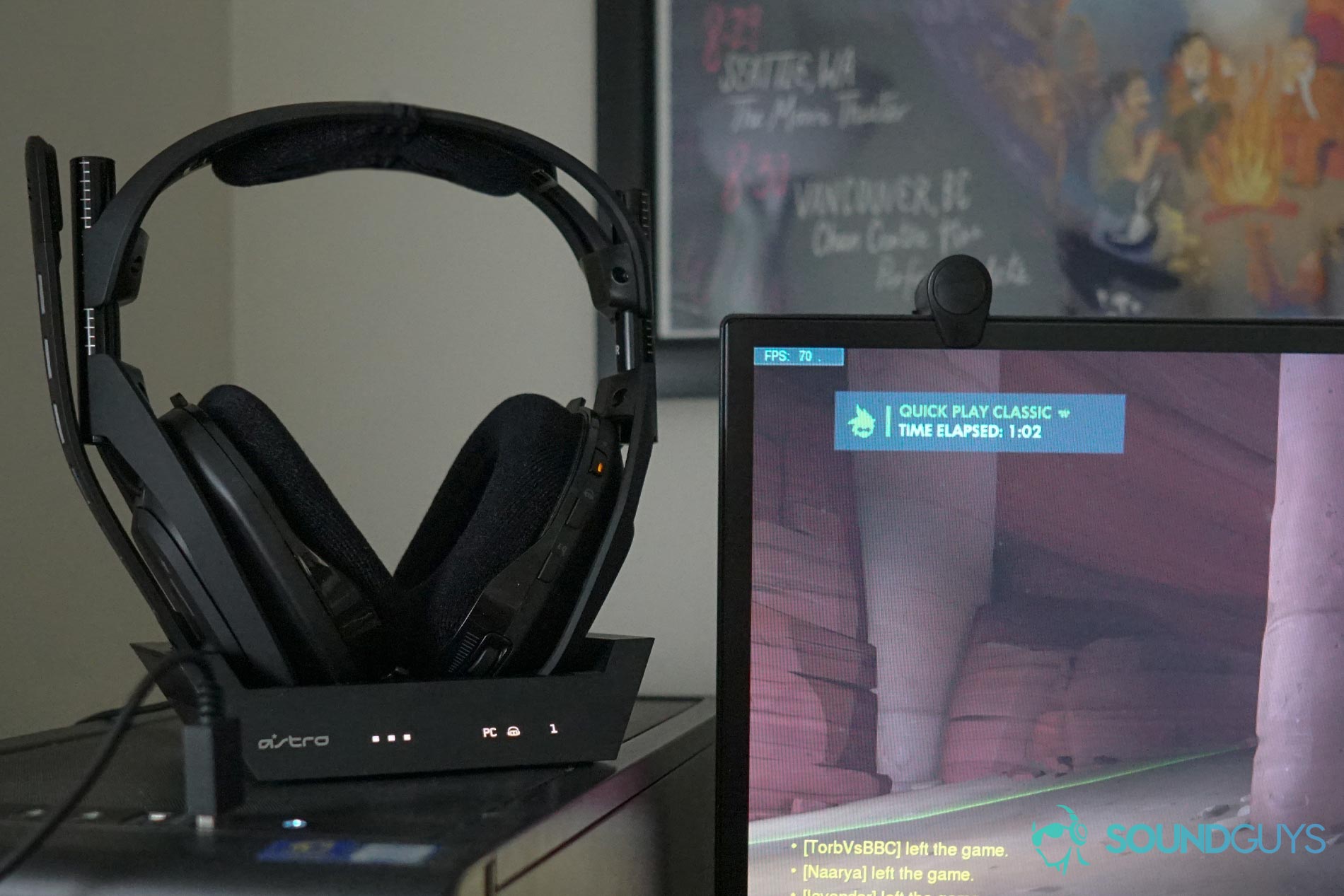 The Astro A50 Wireless sits on its dock on top of a PC next to a monitor running Overwatch in front of a poster for My Brother, My Brother, and Me.