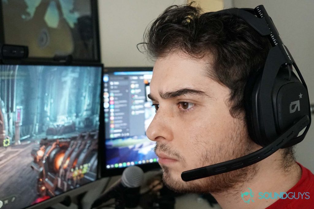 A man wears the Astro A50 sitting at a computer running Doom Eternal and Spotify, with a Shure SM58 microphone in the fore, all in front of an Adventure Zone poster.