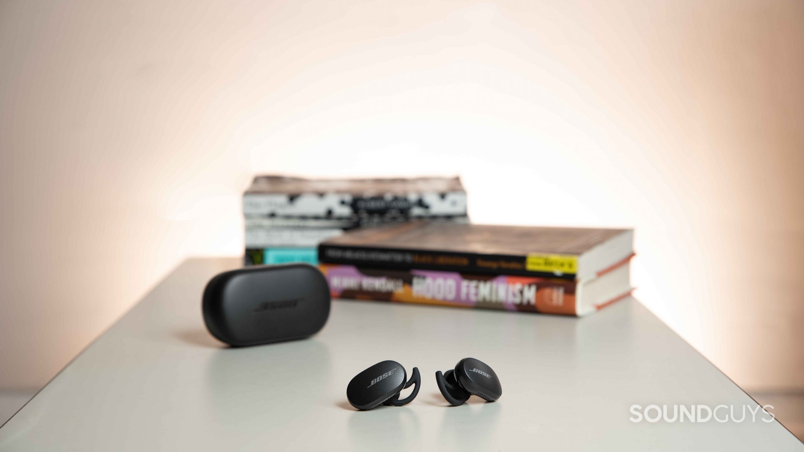 The Bose QuietComfort Earbuds noise canceling true wireless earbuds rest outside of the charging case on a table and in front of a stack of books.