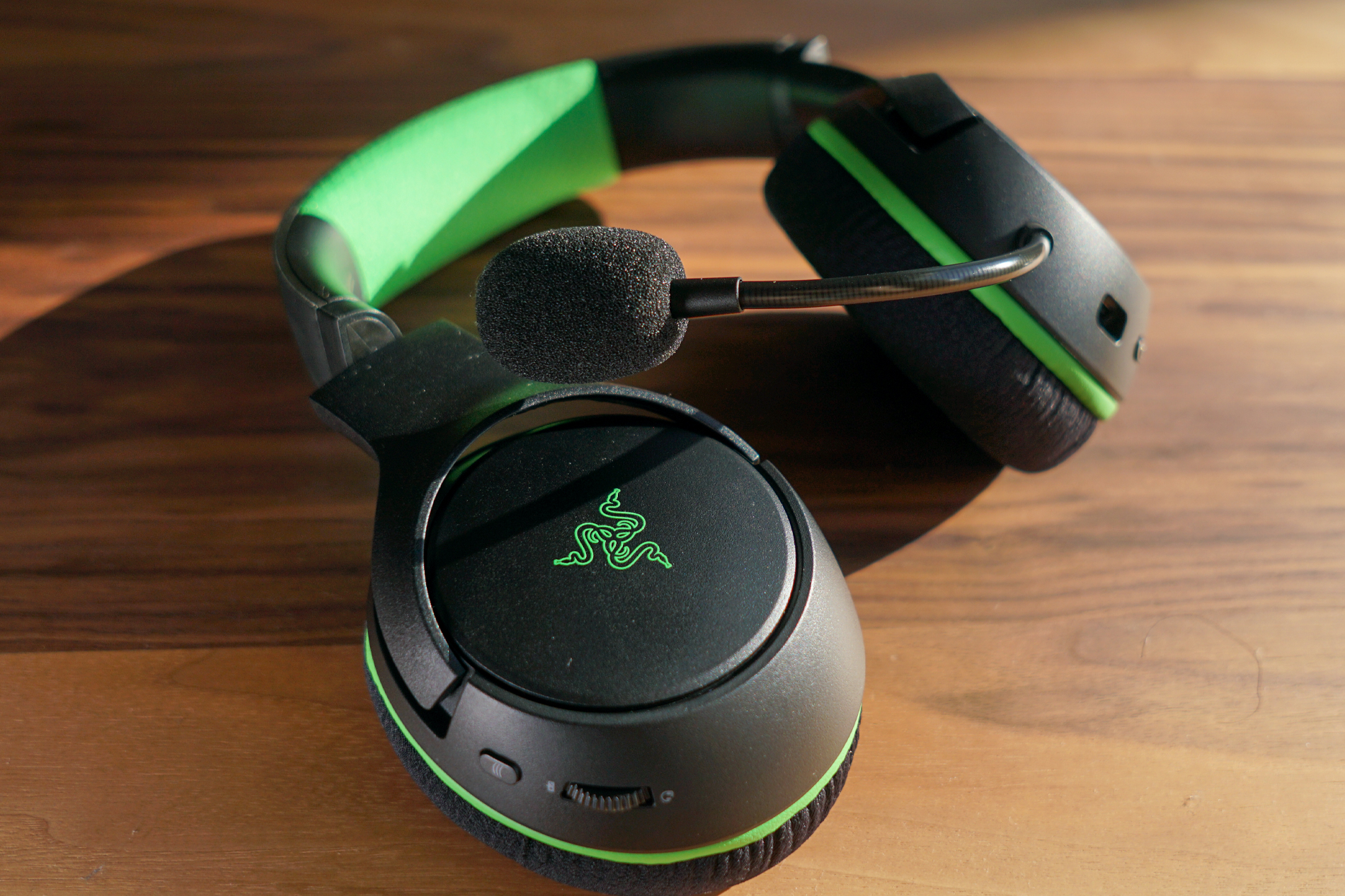 The Razer Kaira Wireless lays on a wooden table with one headphone turned up and the other laying flat.
