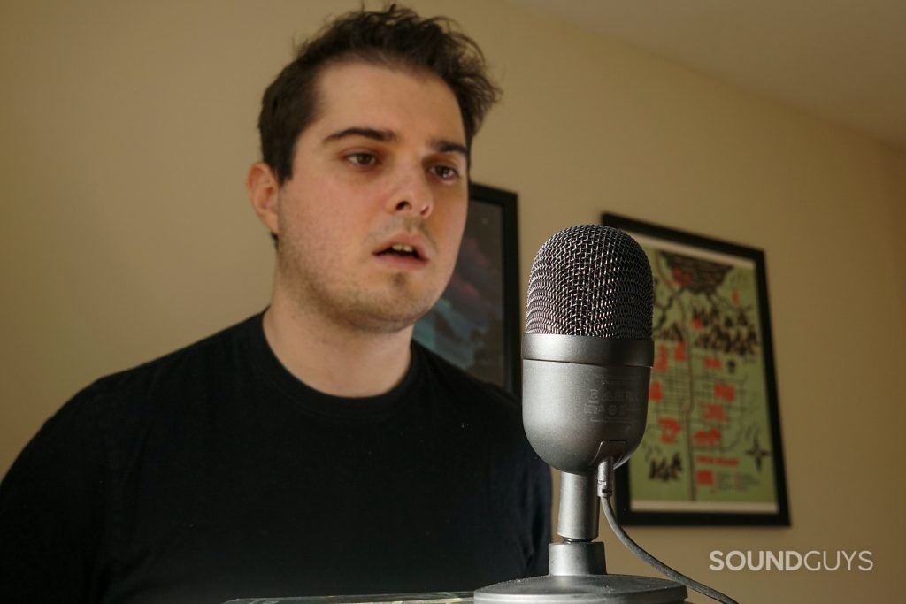 Man sitting behind the Razer Seiren Mini, speaking into it. Paintings hung on the wall are visible behind him.