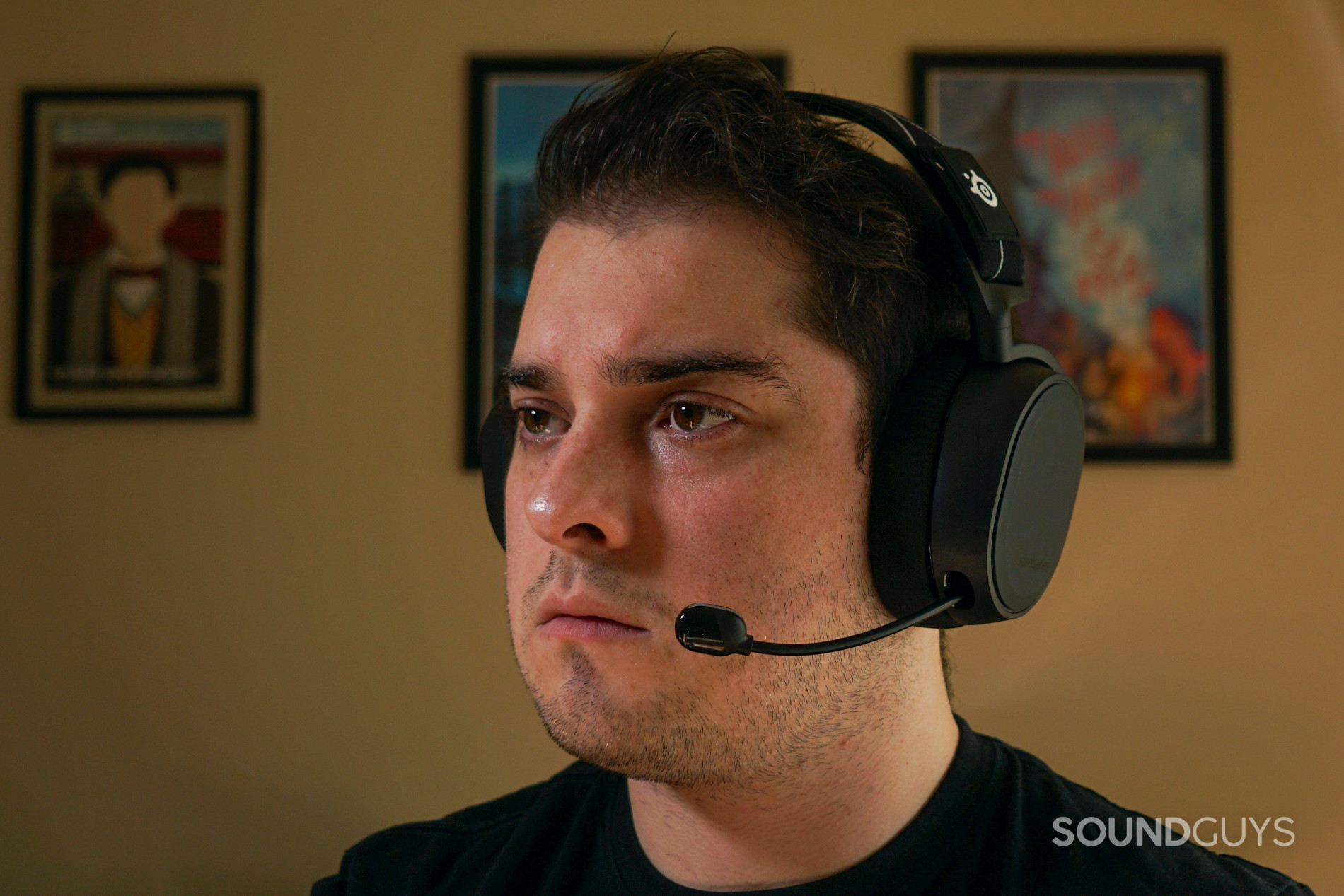 A man wears the SteelSeries Arctis 9 gaming headset in front of posters for My Brother, My Brother and Me, and Canadian Heritage Minutes.