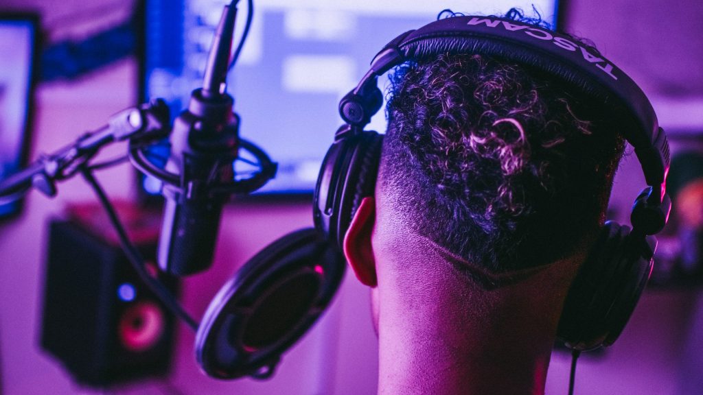 The back of a person's head wearing headphones positioned in front of a recording microphone. In the background the interface of a DAW is visible.