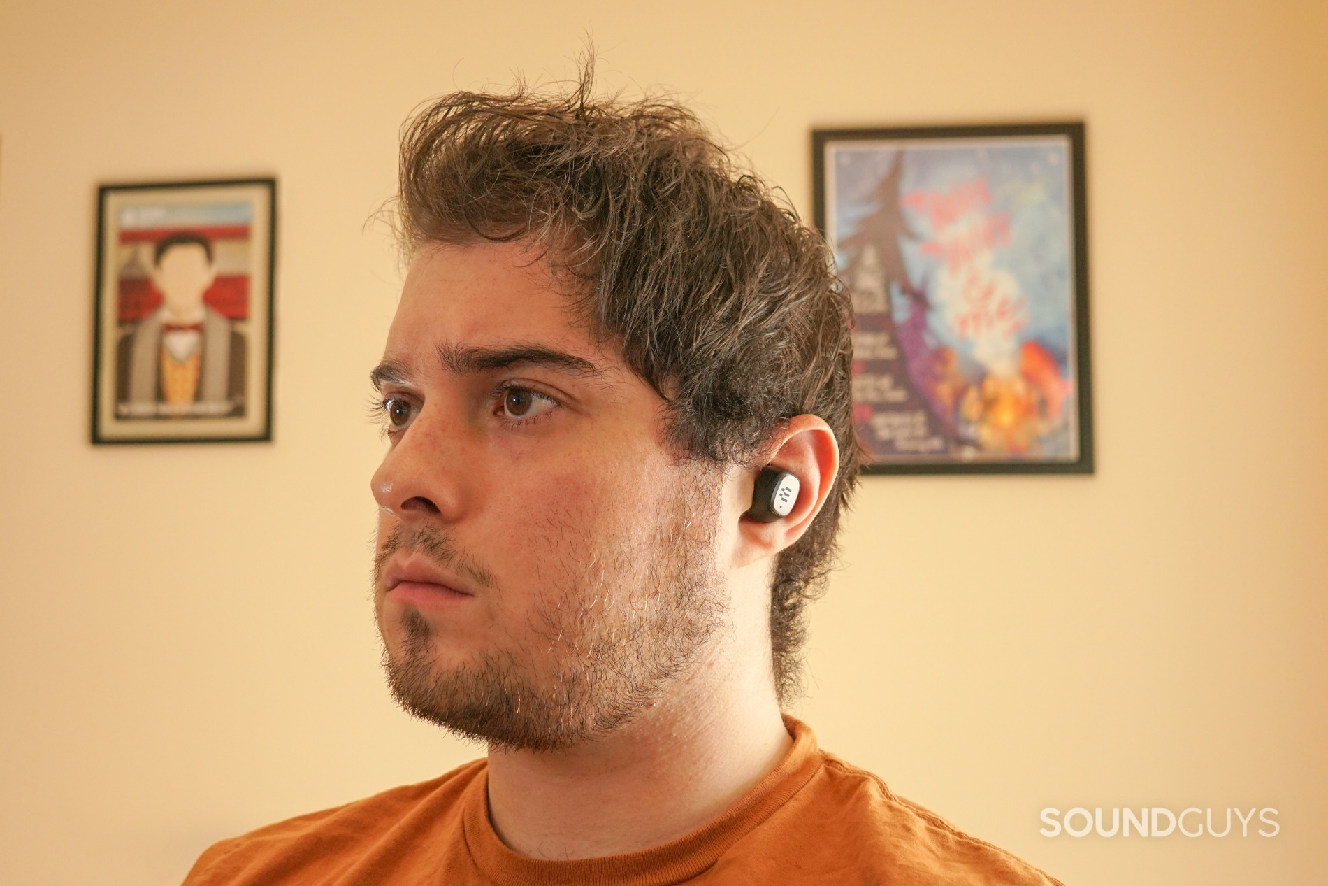 A man wears the EPOS GTW 270 Hybrid true wireless earbuds sitting at a PC, with posters for My Brother, My Brother and Me and Canada Heritage minutes in the background.
