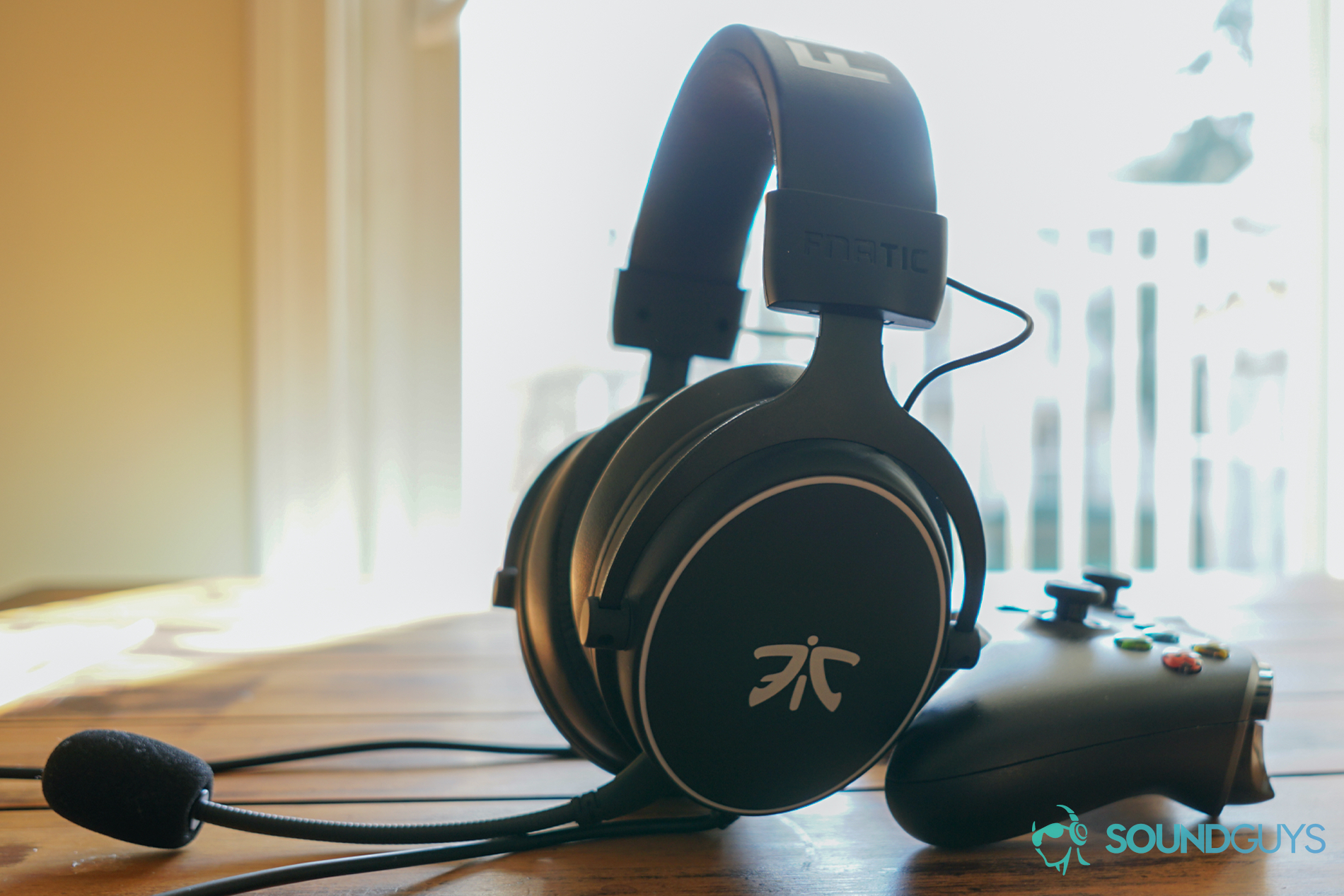 The fnatic react gaming headset sits on a wooden table by a window leaning on an Xbox One controller.