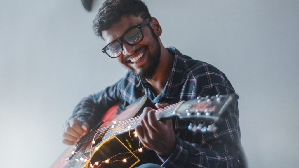 A man smiles while he plays guitar in an empty room.