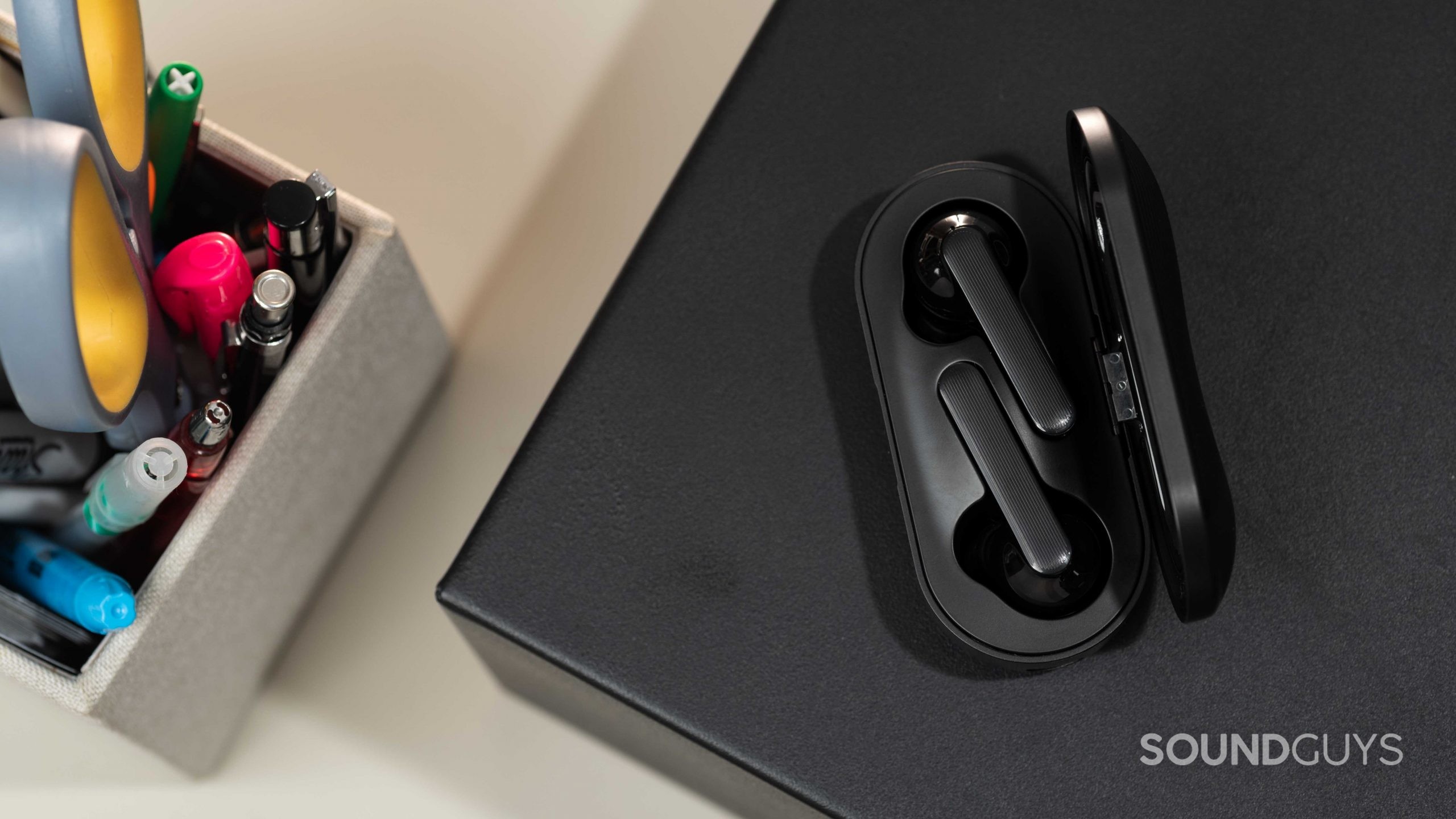 The Mobvoi Earbuds Gesture true wireless earbuds rest inside the USB-C charging case next to a pen holder full of colorful pens and office tools.
