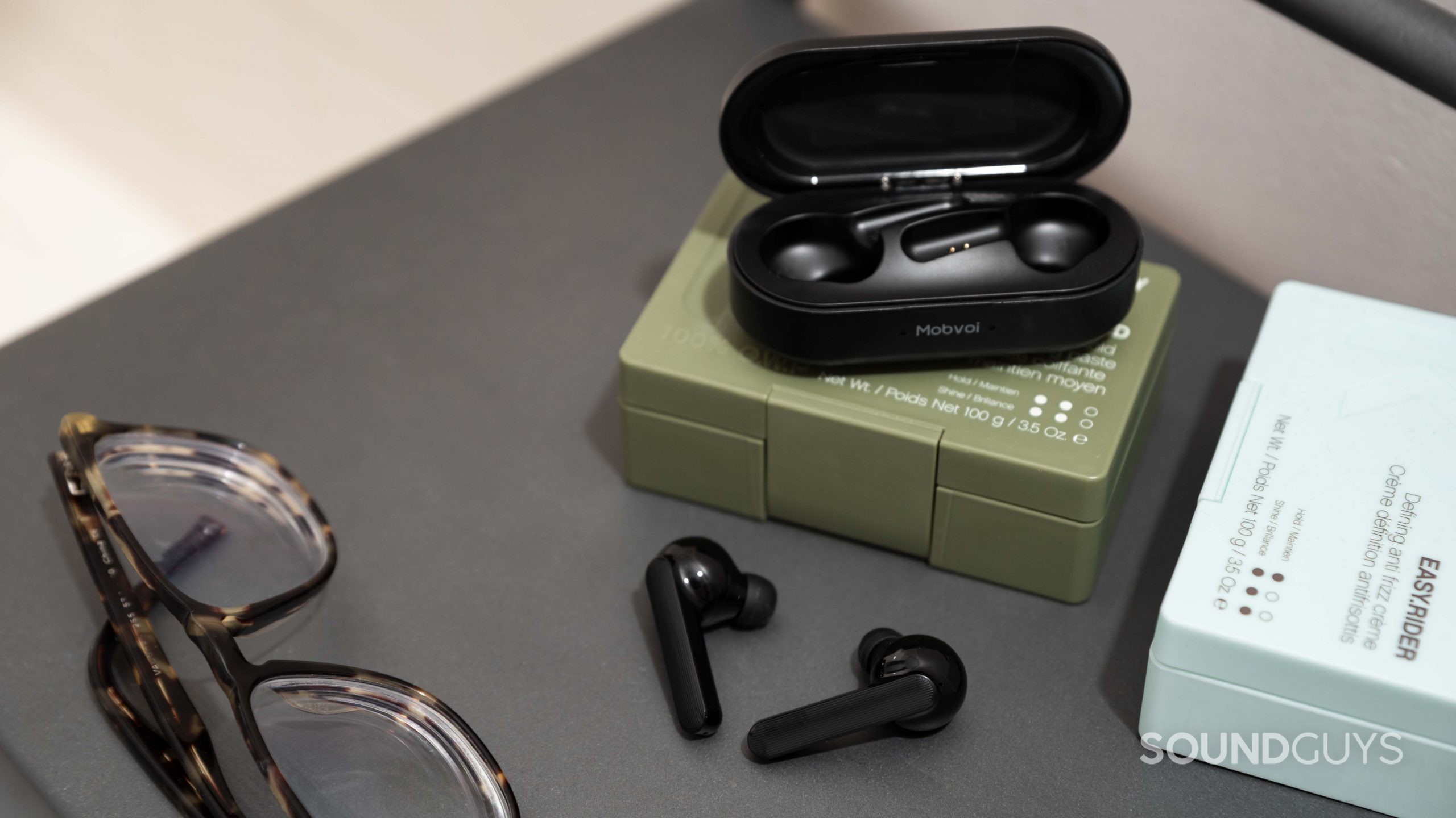 The Mobvoi Earbuds Gesture true wireless earbuds outside of the open USB-C charging case, which rests on top of hair pomade.