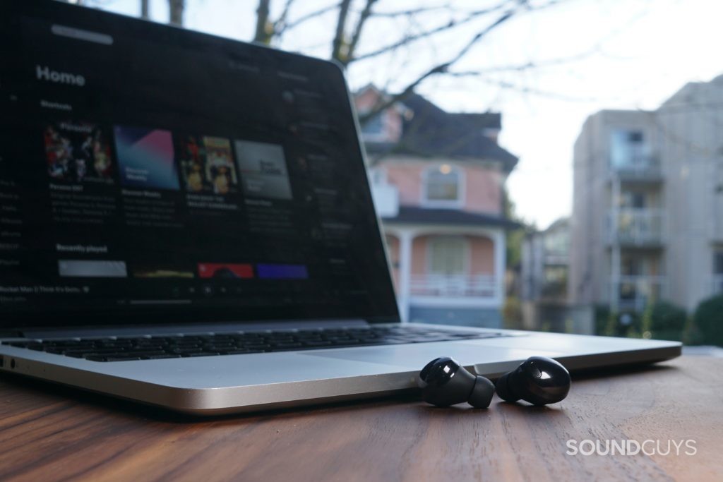The Samsung Galaxy Buds Pro lay on a wooden table in front of a MacBook Pro by a window.