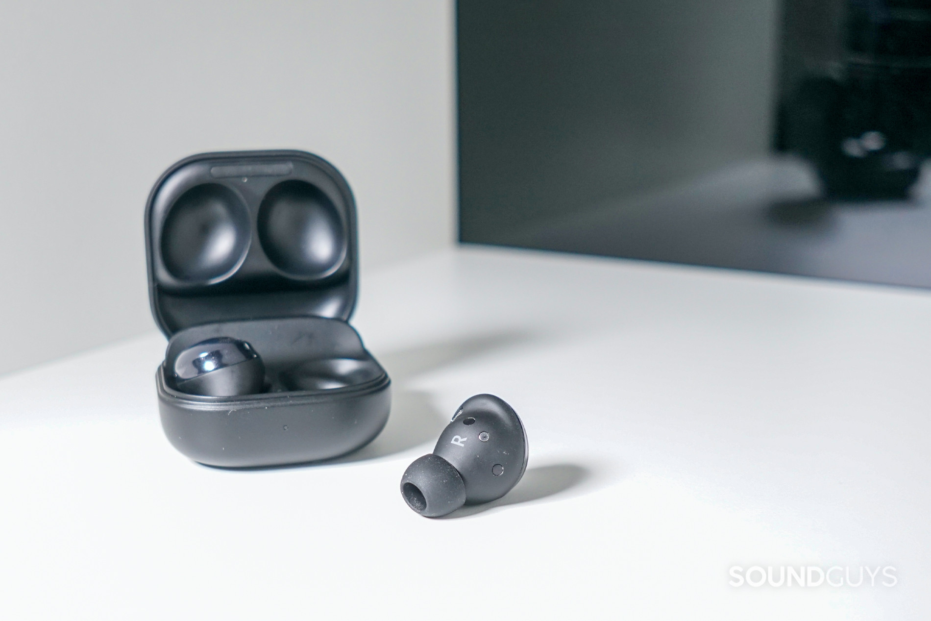 The Samsung Galaxy Buds Pro true Wireless earbuds sit on a white shelf in front of a reflective black surface, with one earbud in the case.