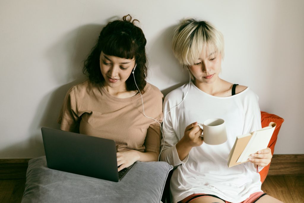 Two women sitting against a wall sharing a pair of wired earbuds.