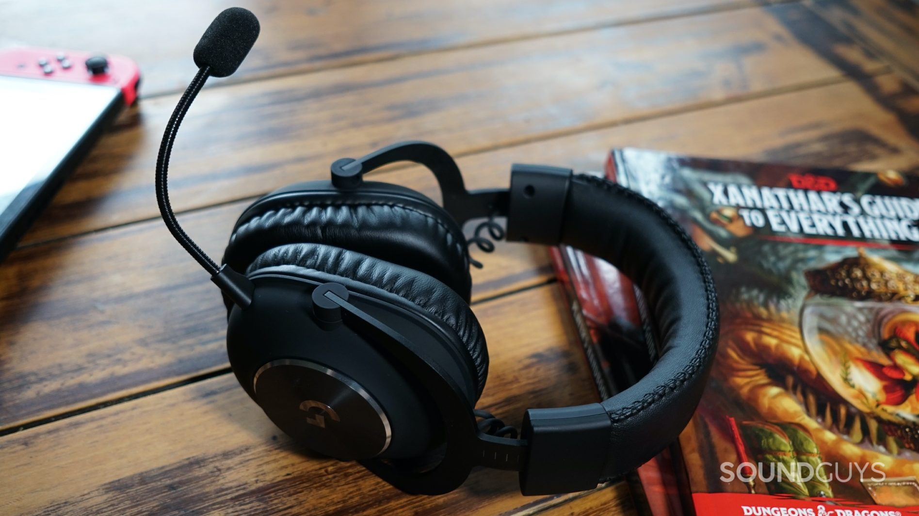 The Logitech G Pro X gaming headset lays on a wooden table leaning on a copy of Xanathar's Guide to Everything, with a Nintendo Switch in the background.