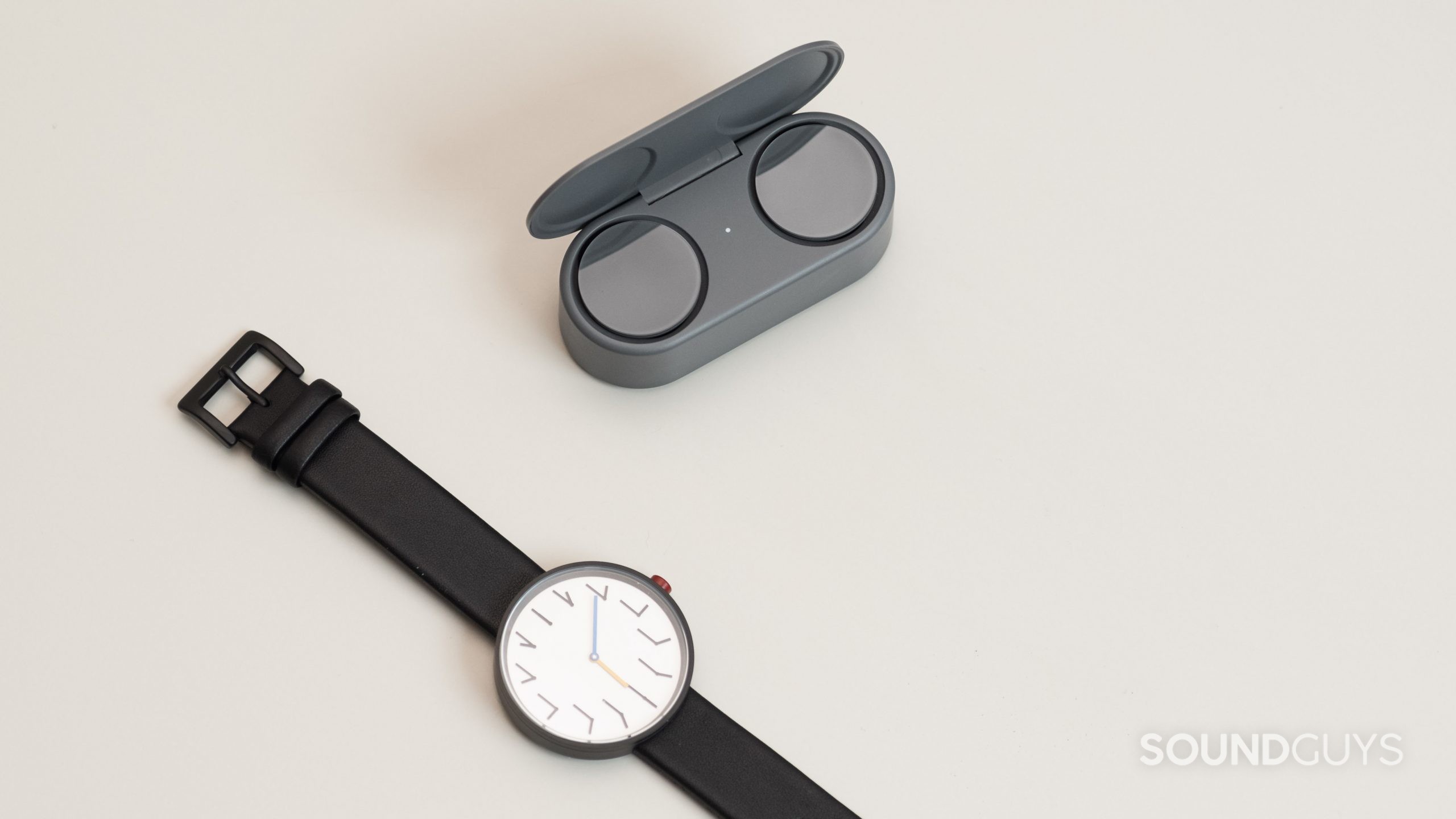 The Microsoft Surface Earbuds open case next to a watch on an off-white surface.