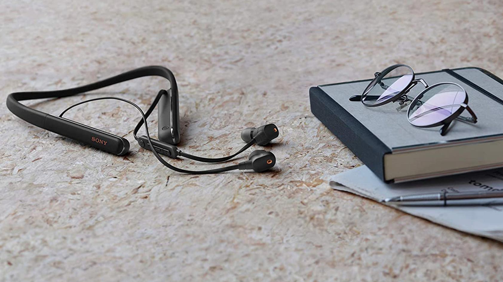 The Sony WI-1000XM2 rest on a surface next to a book and eyeglasses.