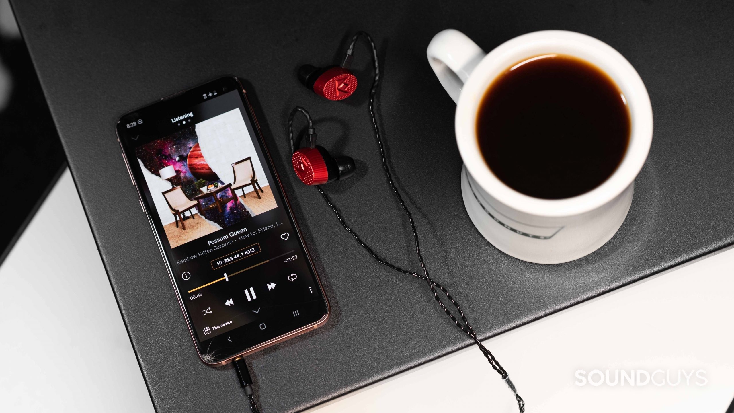 The Qobuz music streaming service app's Listening tab open on a Samsung Galaxy S10e smartphone next to a pair of Massdrop x Noble Kaiser 10 IEMs and a cup of coffee.
