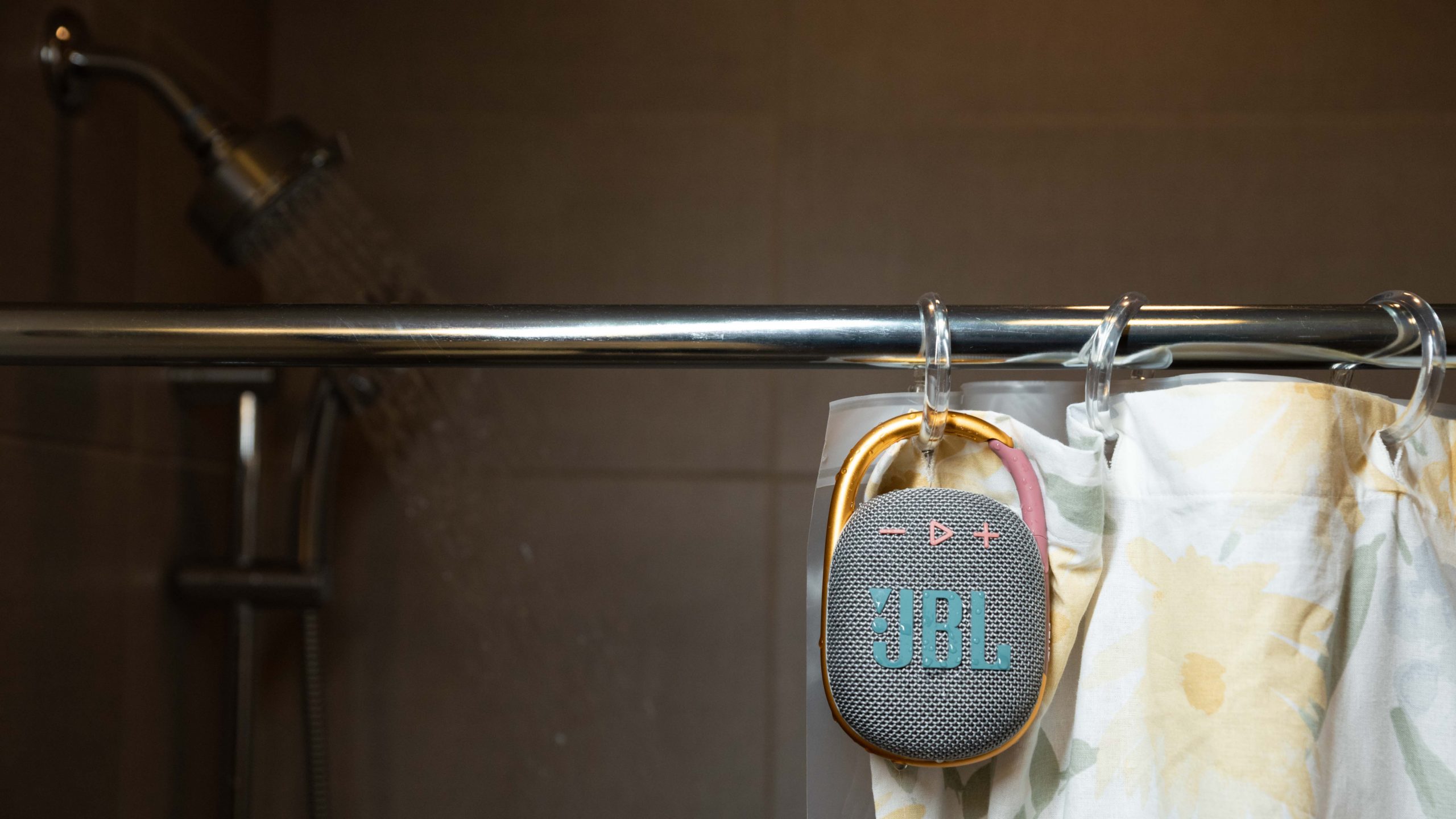 The JBL Clip 4 Bluetooth speaker hands from a shower rod in front of a shower curtain and running shower head.