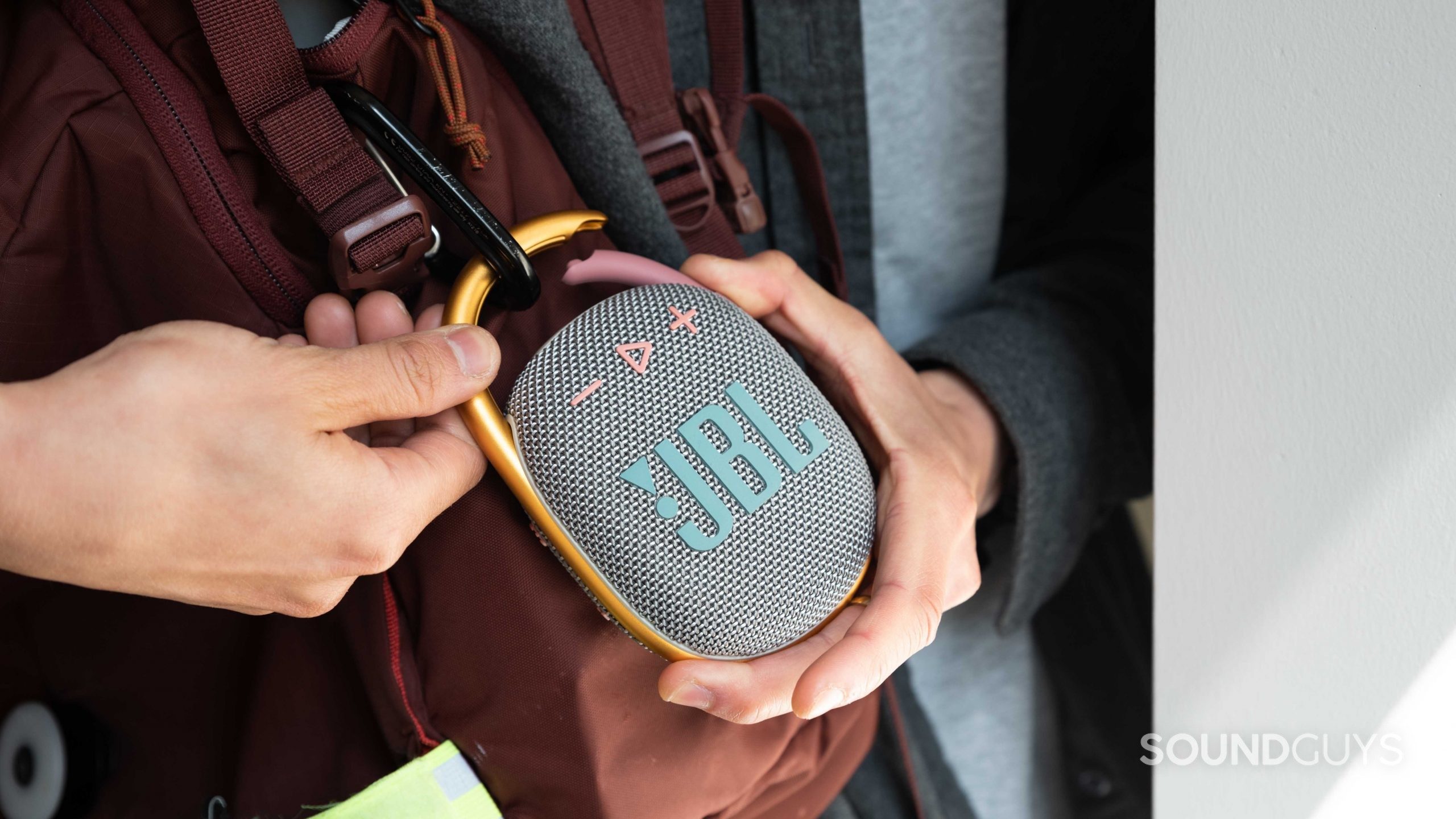 JBL Clip 4_portable Bluetooth speaker-1 Two hands attach the JBL Clip 4 Bluetooth speaker to a red backpack.