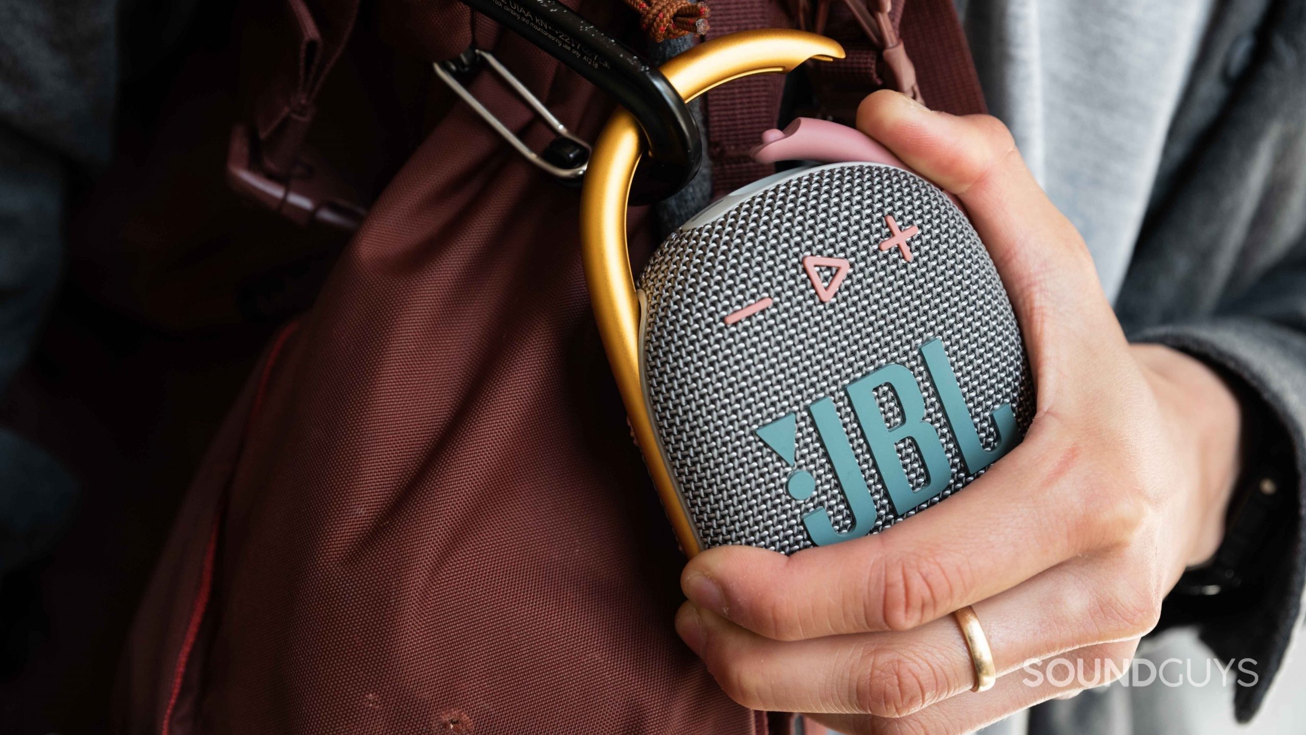 A hand pushes the JBL Clip 4 carabiner down while attaching the speaker to a bag.