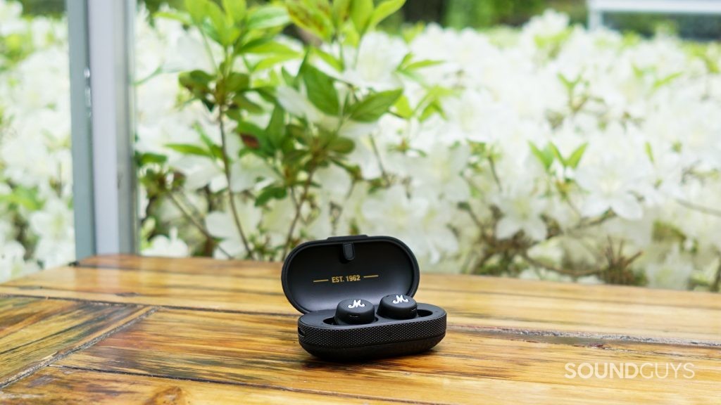 The Marshall Mode II true wireless earbuds sit in their charging case on a wooden table next to an azalea plant
