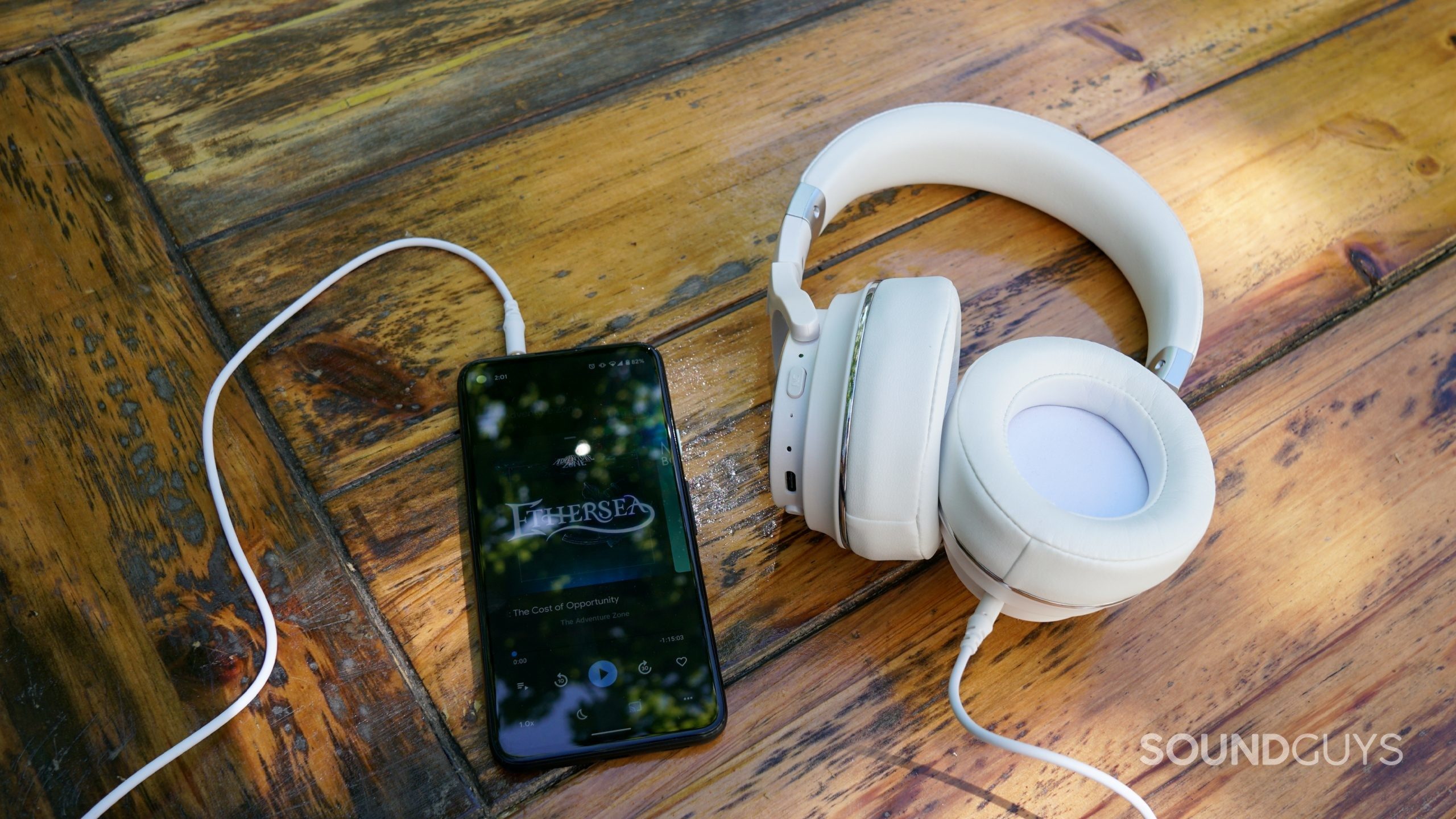 The Raycon Everyday headphones lay on a wooden table connected over 3.5mm to a Google Pixel 4a playing The Adventure Zone
