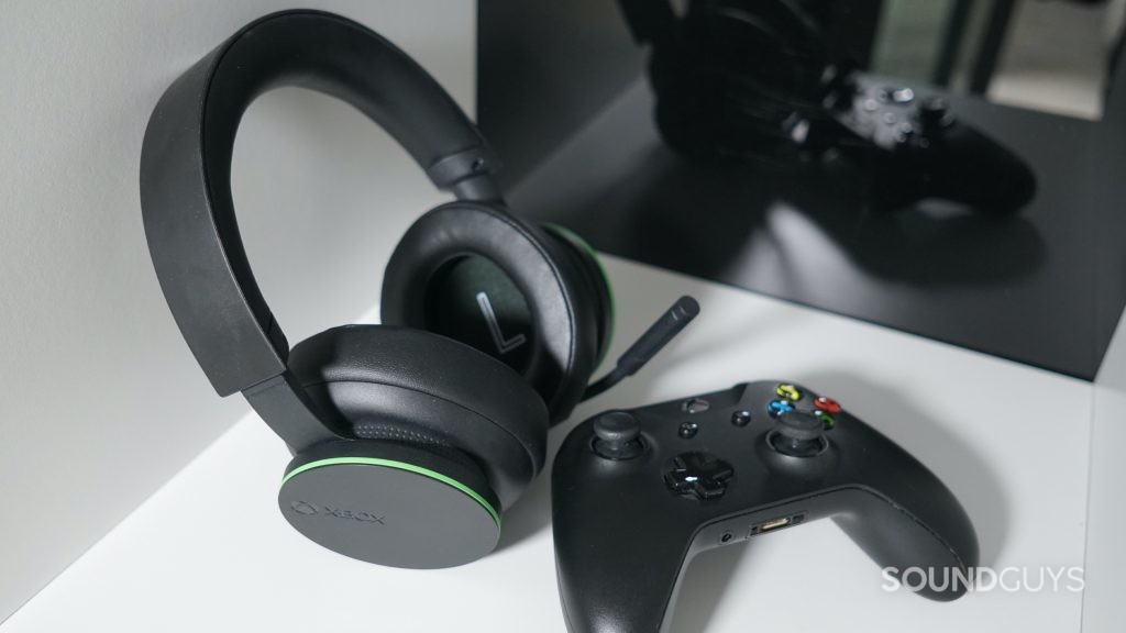 the Microsoft Xbox Wireless Headset lays on a white shelf in front of a black reflective surface, next to an Xbox One controller.