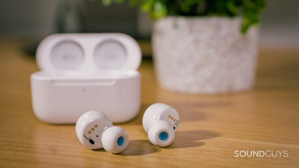 The Amazon Echo Buds 2nd Generation earphones sitting on a wooden desk, with case and plant in background.