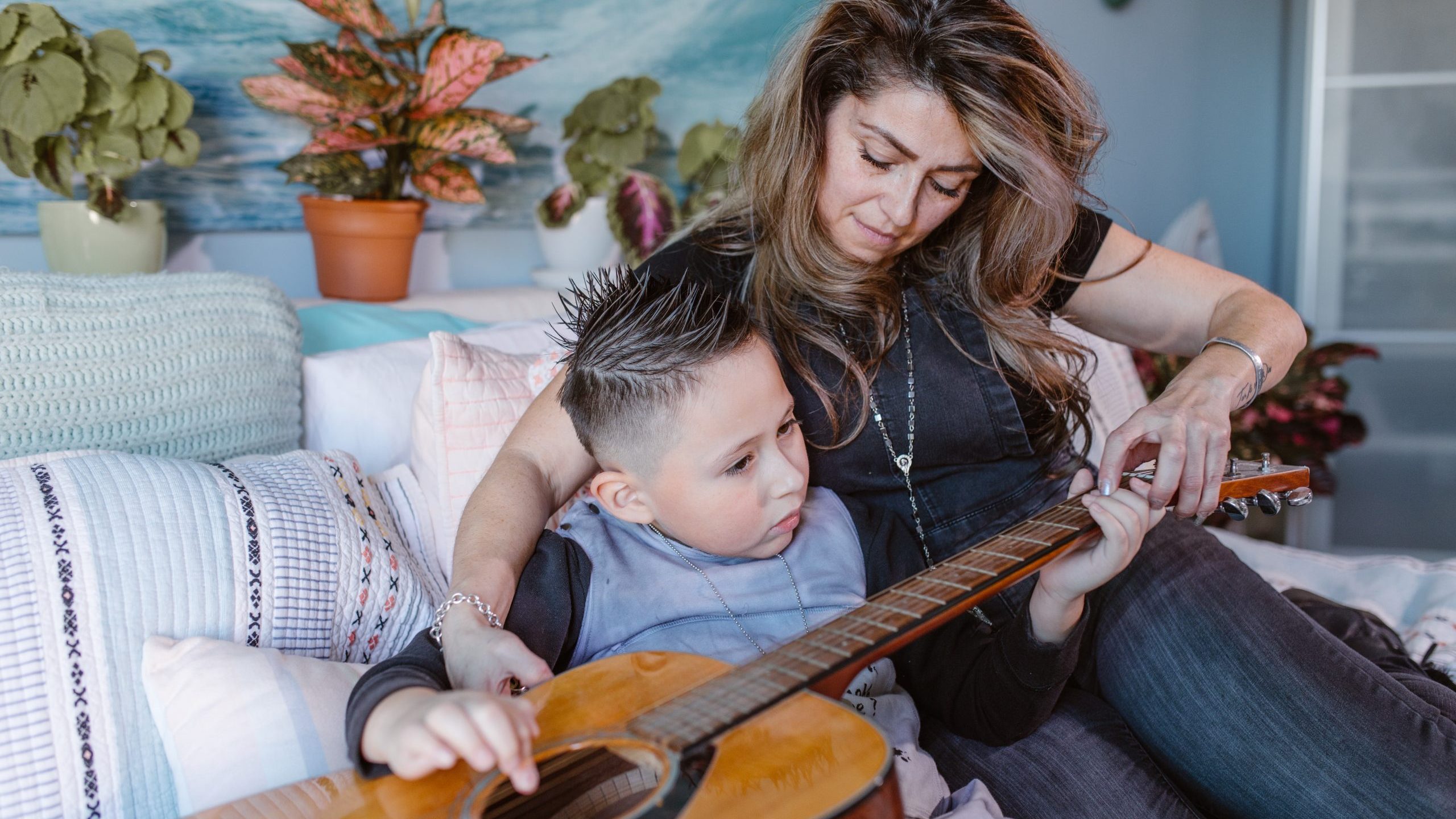 Woman helping young boy play an acoustic guitar.
