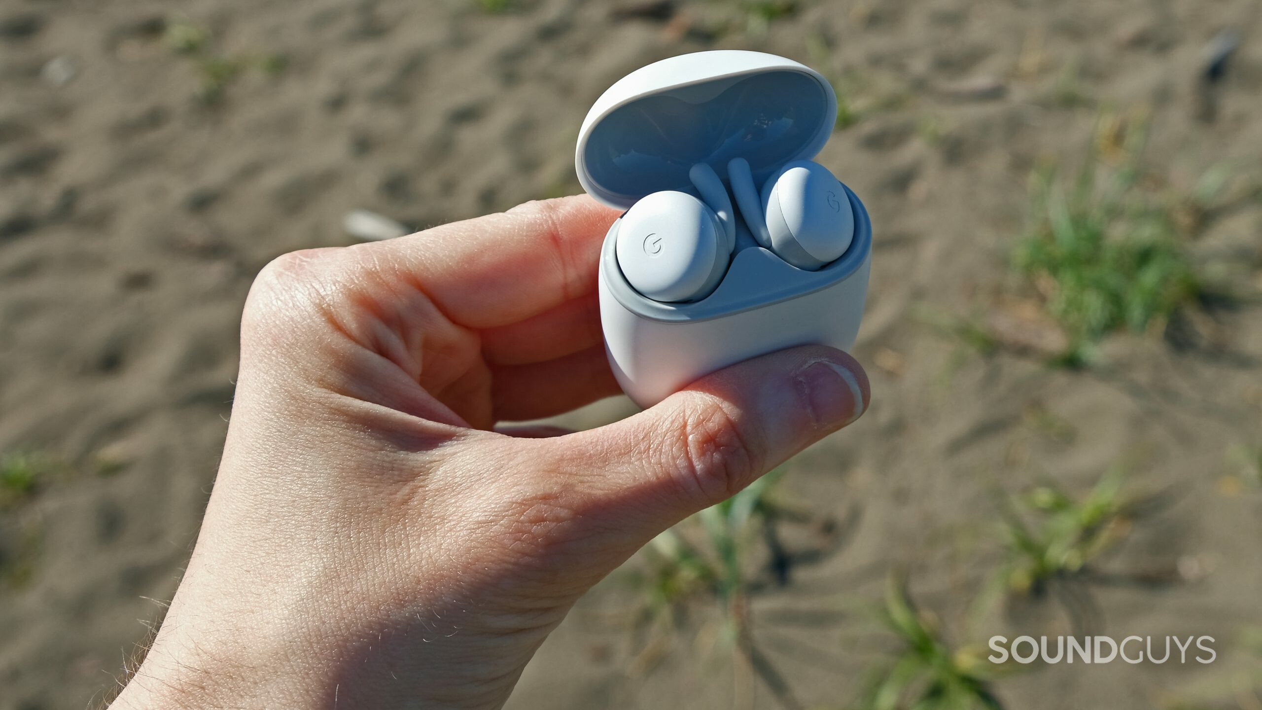 A hand holds the open case of the Google Pixel Buds A-Series in front of a beach.