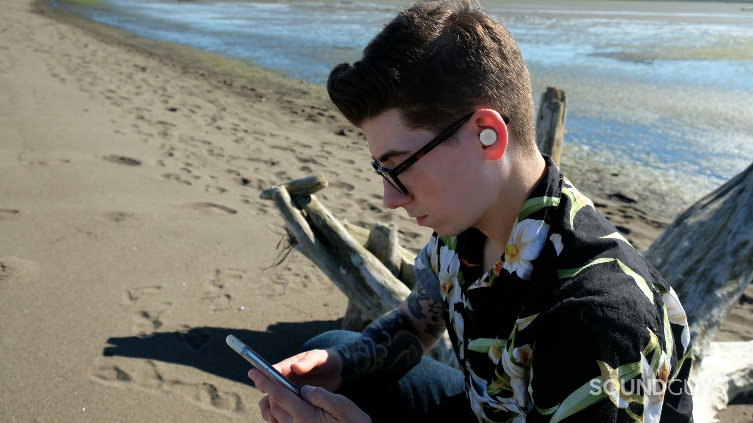 A man is seated at the beach with a smartphone and wearing Google Pixel Buds A-Series.