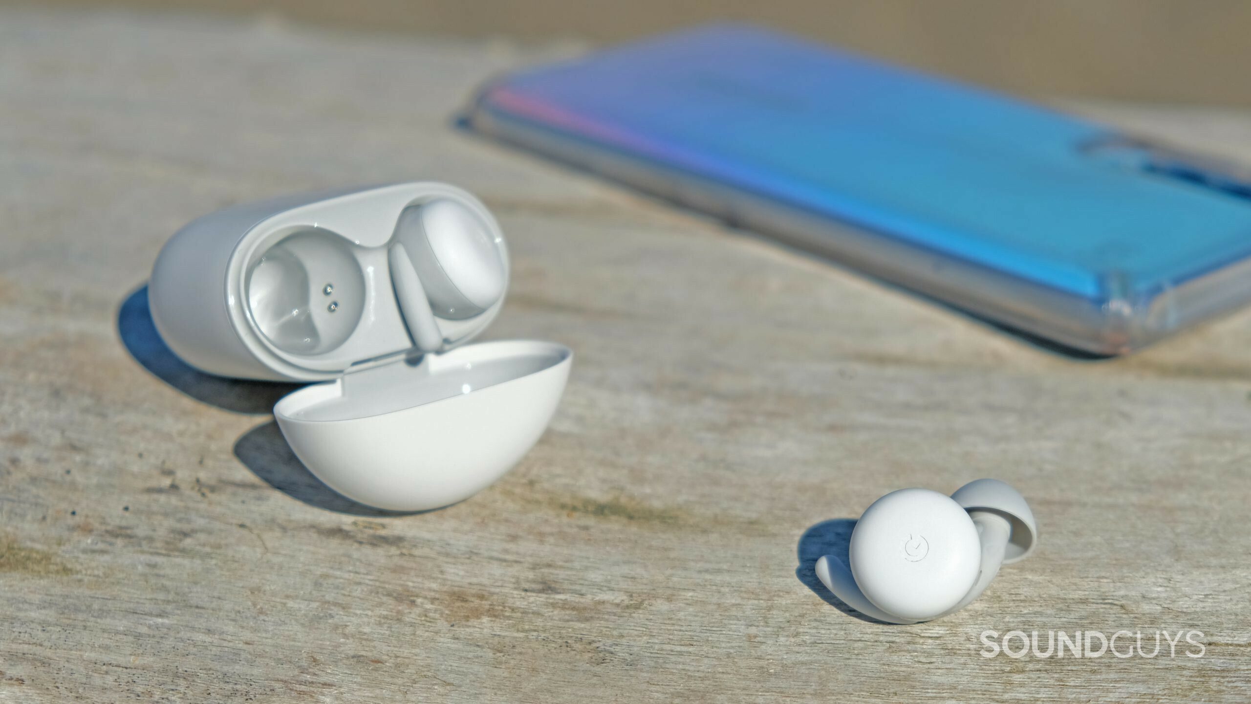 Here's a close up of the Google Pixel Buds A-Series on driftwood with a smartphone.