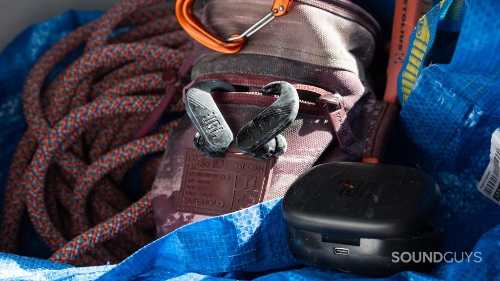 The JBL ENDURANCE PEAK II true wireless workout earbuds hang from a rock climbing chalk bag behind its open charging case.