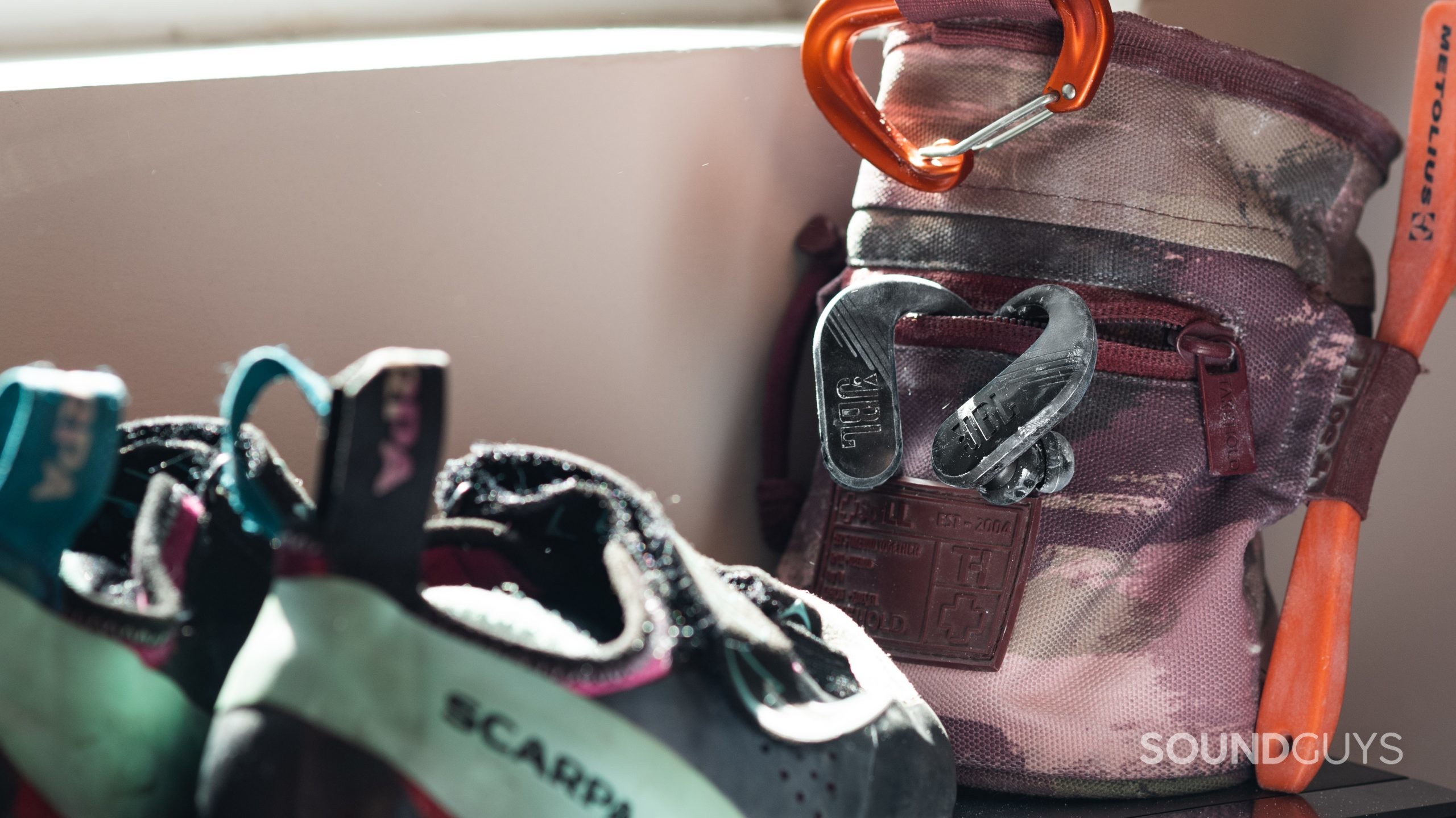 The JBL ENDURANCE PEAK II true wireless workout earbuds hang out of a rock climbing chalk bag behind a pair of rock climbing shoes.