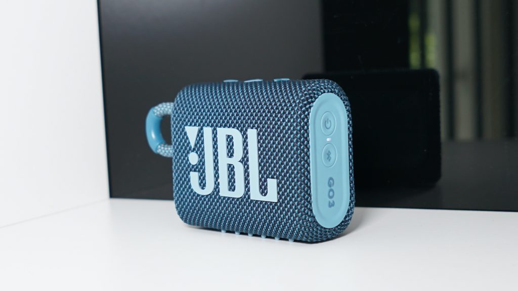 The JBL GO 3 stands on a white shelf in front of a reflective surface.