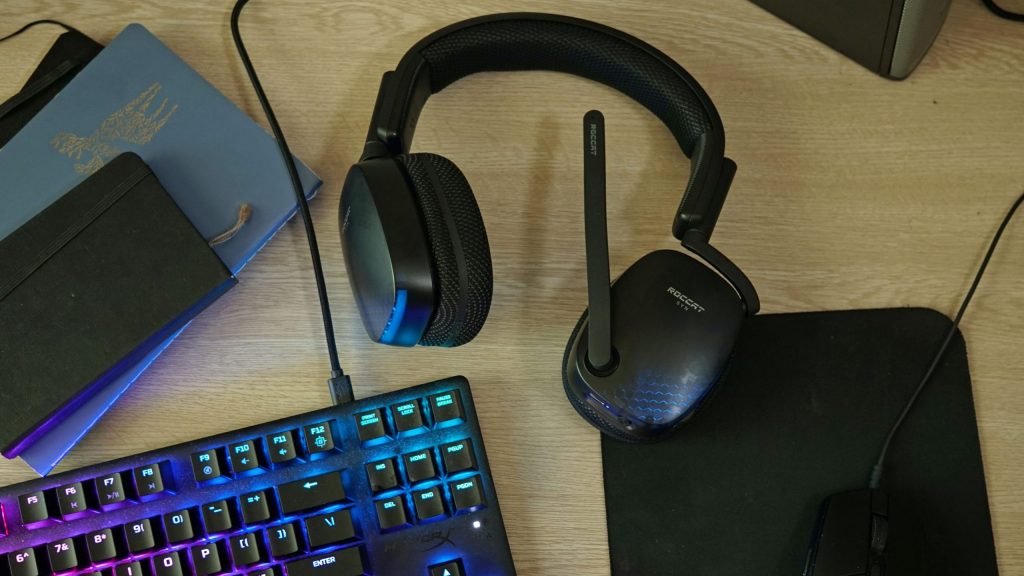 The Roccat Syn Pro Air gaming headset lays on a desk, near assorted notebooks, HyperX mechanical gaming keyboard, and a logitech gaming mouse.