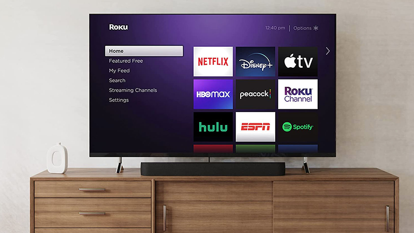 The Roku Streambar Pro on a TV stand with a TV that displays the Roku home page.