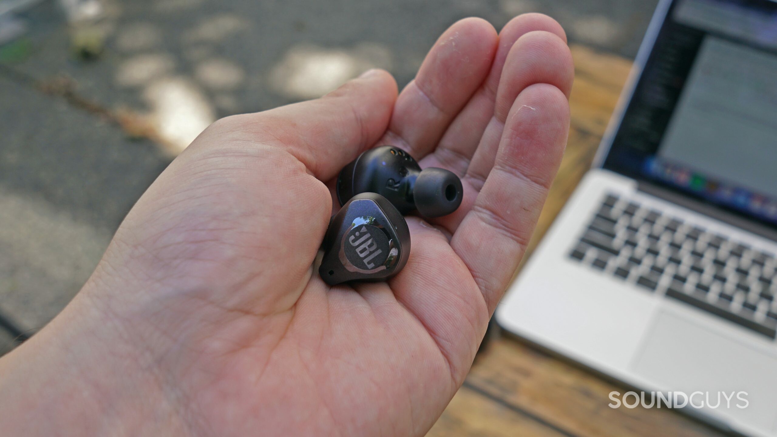 Someone holding the JBL Club Pro Plus earbuds, with an Apple MacBook Pro on a wooden table