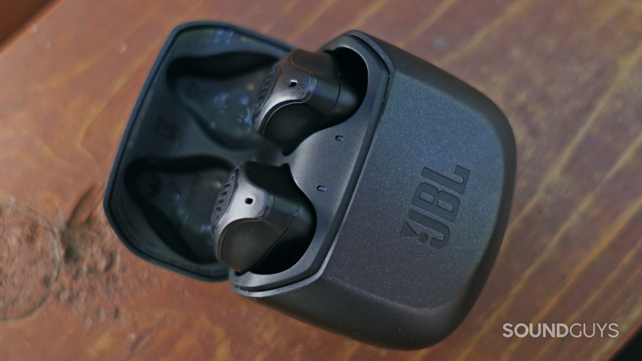 The JBL Club Pro + lays in its charging case on a wooden surface.