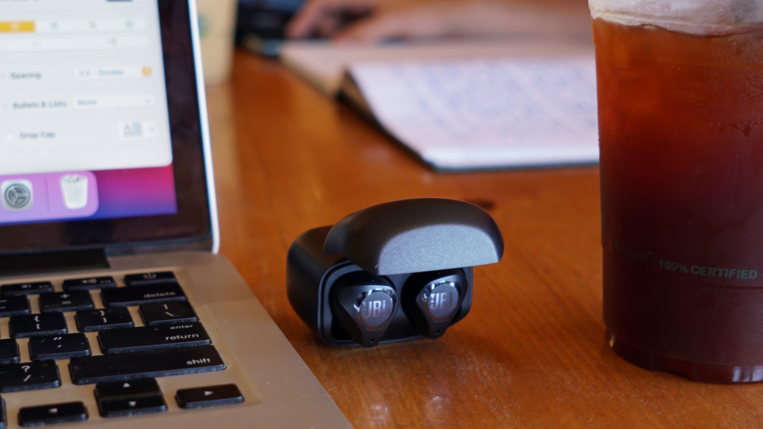 The JBL Club Pro Plus lays inside its case on a cafe table next to a cup of iced coffee and an Apple MacBook Pro.