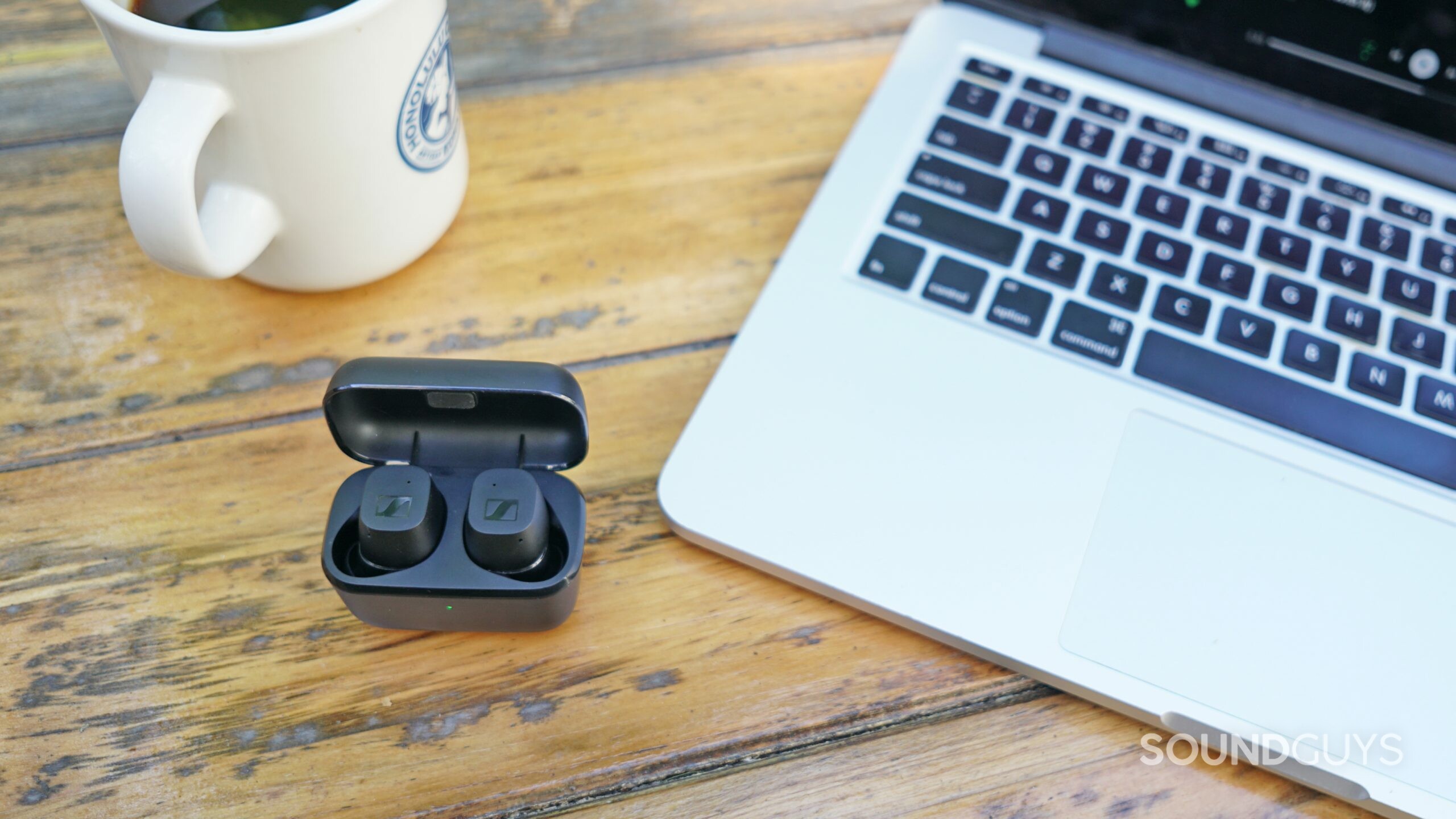 The Sennheiser CX True Wireless sits in its charging case next to an Apple MacBook Pro and a cup of coffee on a wooden table.