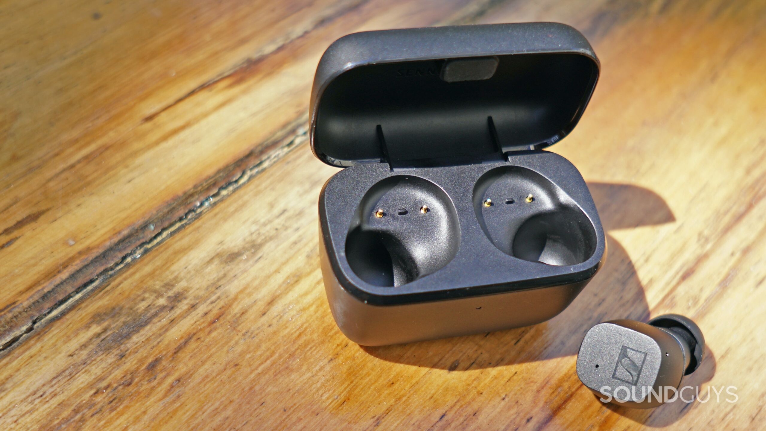 One Sennheiser CX True Wireless earbud sits on a wooden table next to its open charging case.