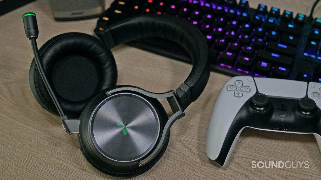 The Corsair Virtuoso RGB Wireless XT lays on a desk next to a PlayStation DualSense controller, in front of a HyperX mechanical gaming keyboard a Bose Companion speaker.