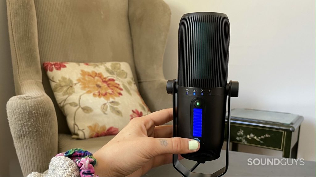 A womans hand using the JLab Talk PRO USB microphone on a table with an armchair in the background.