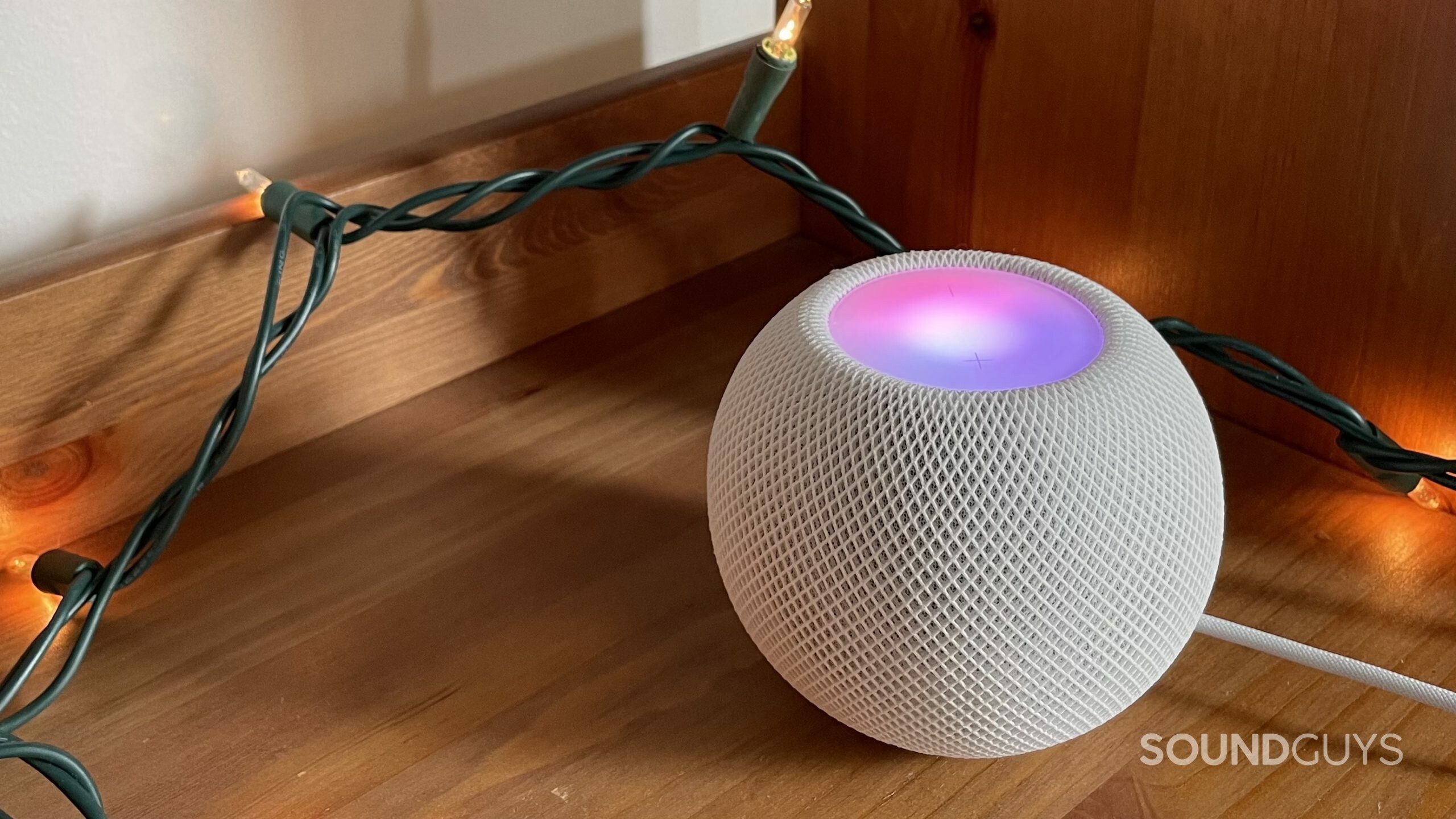Apple HomePod mini with touchpad glowing pink and purple on a bookshelf. A string of lights is used to frame the HomePod mini.