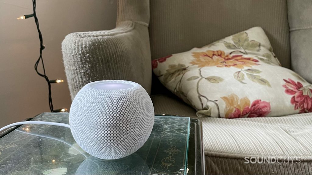 Apple HomePod mini resting on a table with an arm chair and string lights in the background.