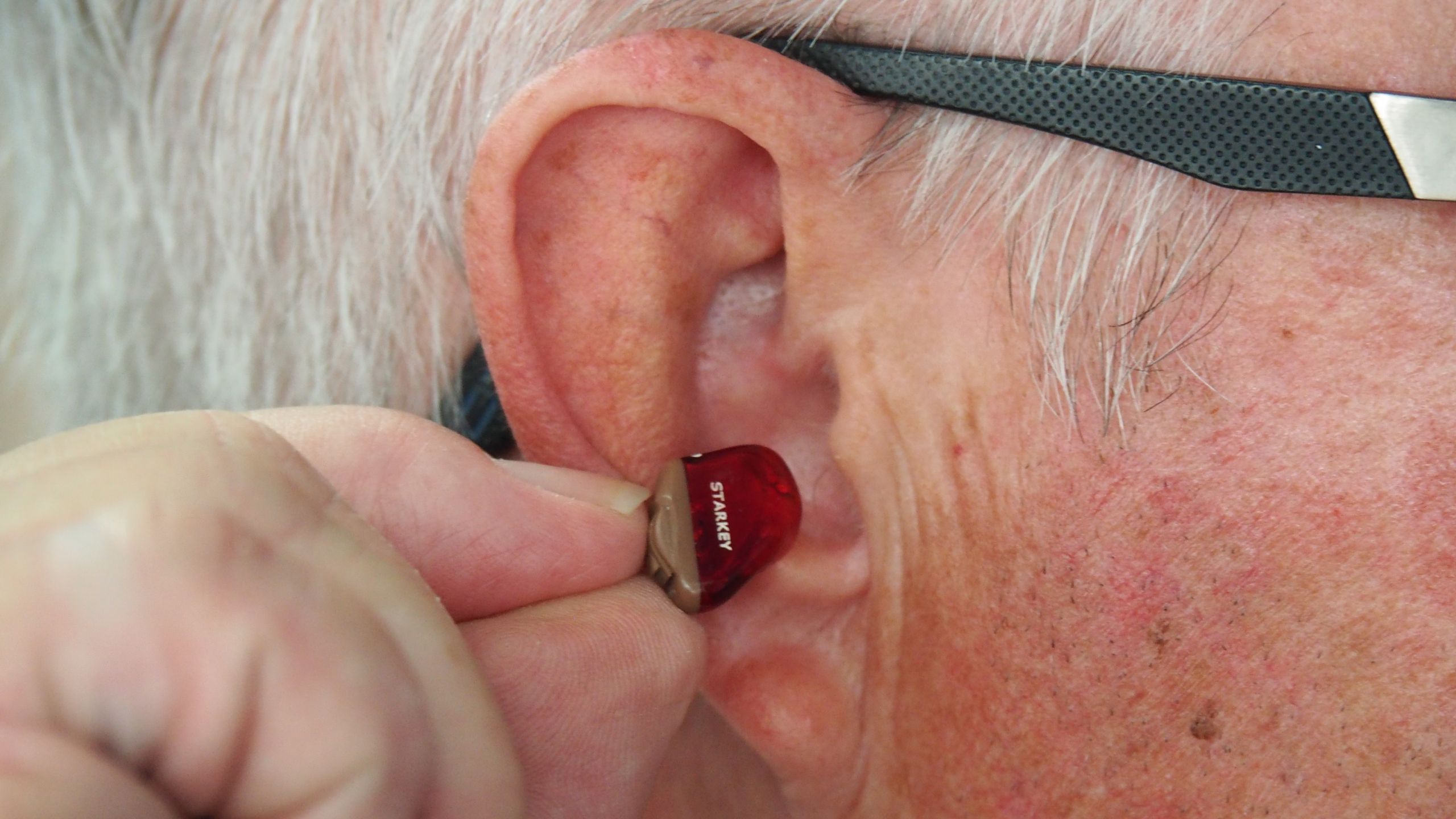 Elderly man with glasses inserting ITC hearing aid into his right ear.